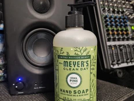 Mrs. Meyer’s Clean Day Iowa Pine hand soap bottle on a studio desk with a speaker and audio equipment in the background.