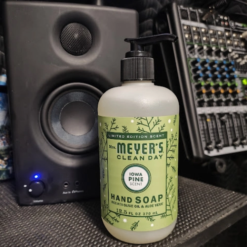 Mrs. Meyer’s Clean Day Iowa Pine hand soap bottle on a studio desk with a speaker and audio equipment in the background.
